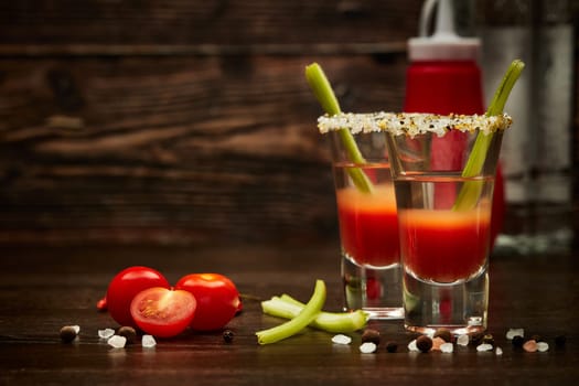 Two shot glasses with cocktail Bloody Mary, celery, spices and ripe tomatoes on dark wooden background with copy space