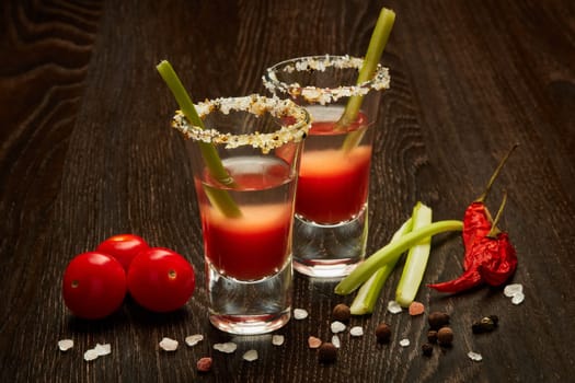 Two shot glasses with cocktail Bloody Mary, celery, spices and ripe tomatoes on dark wooden background