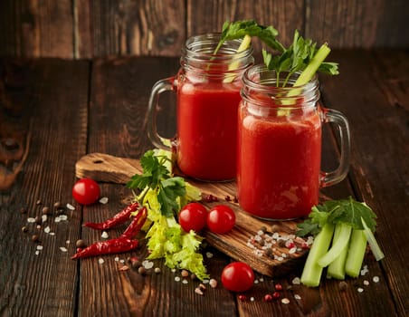 Two glasses with fresh tomato juice, celery, parsley, spices and ripe tomatoes on dark brown wooden background with copy space. Healthy beverage, vegetable juice
