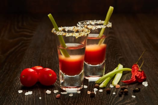 Two shot glasses with cocktail Bloody Mary, celery, spices and ripe tomatoes on dark wooden background