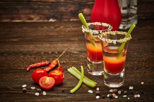 Two shot glasses with cocktail Bloody Mary, celery, spices and ripe tomatoes on dark wooden background with copy space