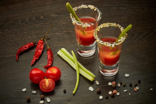 Two shot glasses with cocktail Bloody Mary, celery, spices and ripe tomatoes on dark wooden background. Top view