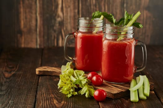 Two glasses with fresh tomato juice, celery, parsley and ripe tomatoes on dark brown wooden background with copy space. Healthy beverage, vegetable juice