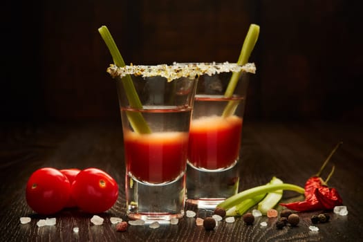Two shot glasses with cocktail Bloody Mary, celery, spices and ripe tomatoes on dark wooden background