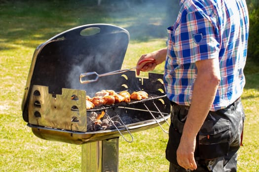 A man at the charcoal grill uses grill tongs to place the already seasoned raw meat on the grill. Selective focus on the grill against a green lawn background on a sunny spring day. BBQ concept. Macro.
