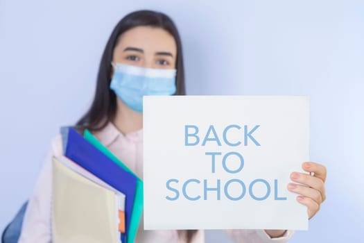 High school girl with mask on her showing back to school message. Student girl ready for school during the coronavirus pandemic. Focus on her hand with paper.
