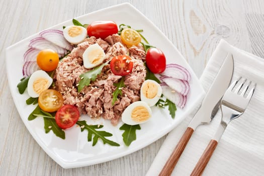 Tuna salad with arugula and tomatoes on white plate on light wooden background. Top view. Healthy food, seafood menu, Setting table
