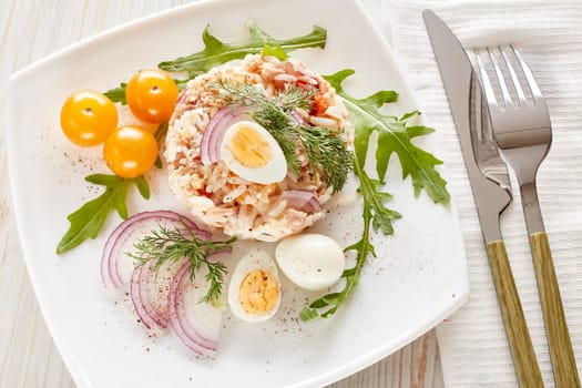Tuna salad with  rice, arugula and tomatoes on white plate on light wooden background. Top view. Healthy food, seafood menu, Setting table