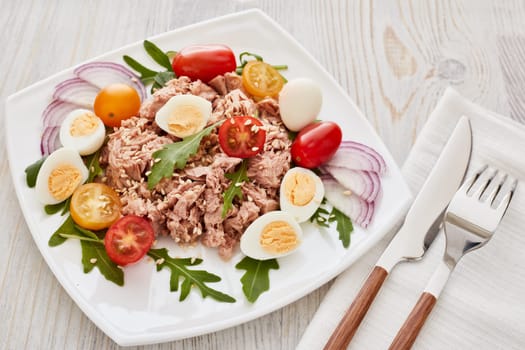 Tuna salad with arugula and tomatoes on white plate on light wooden background. Top view. Healthy food, seafood menu, Setting table