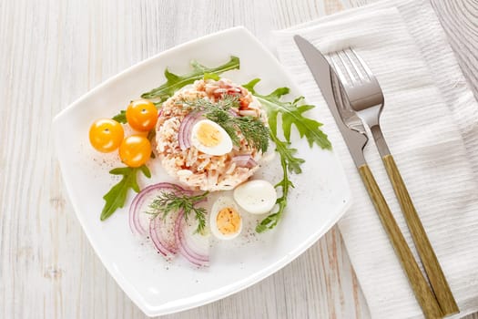 Tuna salad with  rice, arugula and tomatoes on white plate on light wooden background. Top view. Healthy food, seafood menu, Setting table