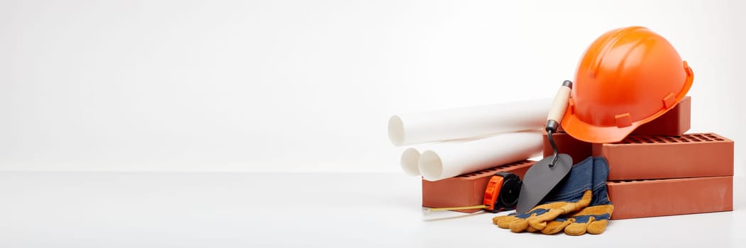 Construction concept. Stack of new bricks with masonry trowel, architectural blueprints, tape measure, construction orange hard hat and protective  gloves on white background with copy space.