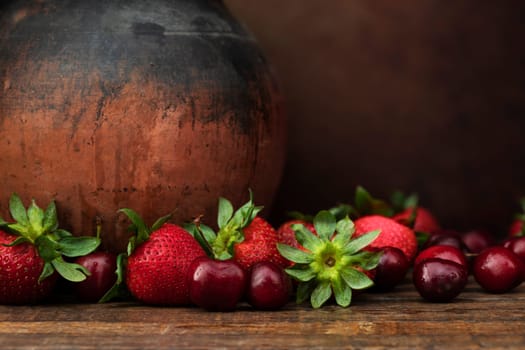 Strawberries and cherries on dark wooden table with rustic pottery jug and copy space.