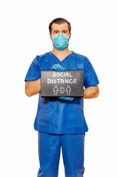 Male doctor in scrubs with protective face mask and gloves holding tablet with chalk text Social Distance. Plain white background. Coronavirus concept