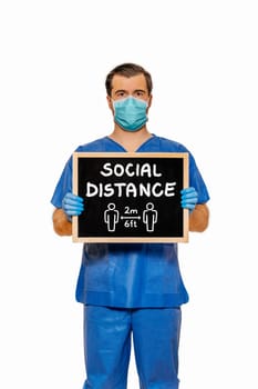 Male doctor in scrubs with protective face mask, holding blackboard with chalk text Social Distance. Plain white background. Coronavirus concept