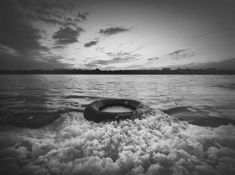 Old vehicle tire thrown on the shore of a pond, covered with fluffy surf foam generated by waves. Scenery sunset view, moody and dramatic background. Environment pollution and ecology concept.