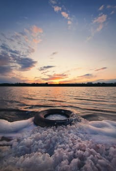 Fine art vertical shot, old vehicle tire thrown on the shore of a pond, covered with fluffy surf foam created by waves. Scenery landscape, peaceful sunset sky background over the city on the horizon.