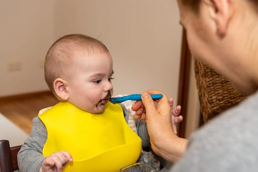 Adorable little baby boy in feeding chair being spoon fed by his mother