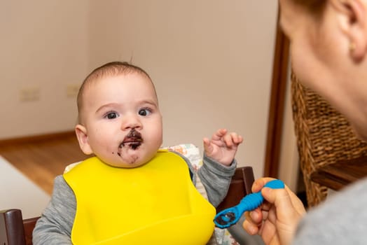 Adorable little baby boy in feeding chair being spoon fed by his mother