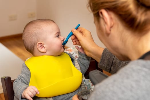 Adorable little baby boy in feeding chair being spoon fed by his mother