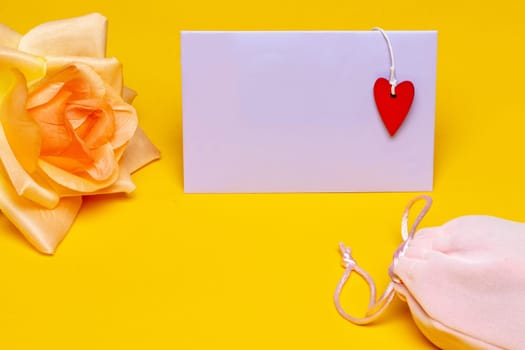 Greeting card template. Close-up of a beautiful yellow blossom of flower and a empty tag with a red heart on it and a pink fabric gift drawstring bag on yellow background. Design template for mother day, wedding, valentine, birthday or other holidays. Macro. Space.
