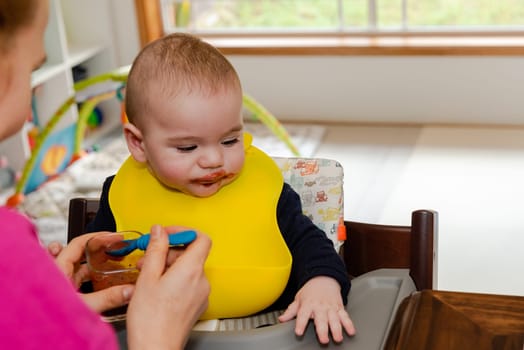 Adorable little baby boy in feeding chair being spoon fed by his mother