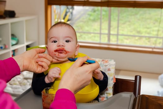 Adorable little baby boy in feeding chair being spoon fed by his mother
