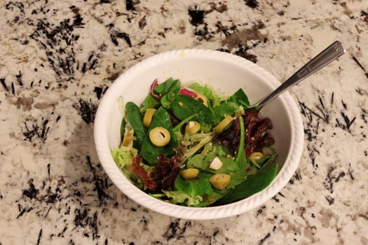Fresh spring mix salad with balsamic vinegar dressing along with garlic stuffed olives. Variety of organic lettuce leaves with balsamic vinegar in a bowl on a kitchen counter with a fork close-up.