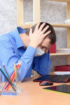 A man sits at a desk holding his head in a medical mask