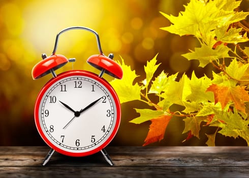 Vintage red alarm clock against the background of yellow autumn maple leaves. Return to wintertime. The old clock next to autumn leaves on a wooden table in front blurred background with copy space.