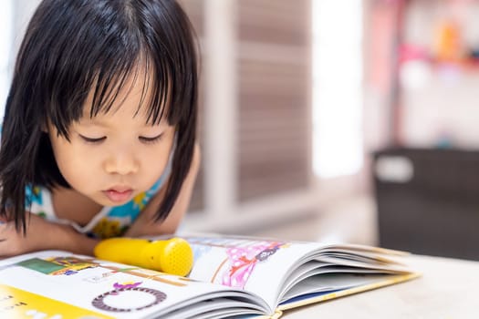 Asian girl child reading interactive book in living room at home as home schooling while city lockdown because of covid-19 pandemic across the world. Home Scholling prepare for Preschool concept.
