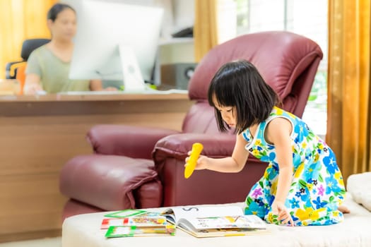 Asian girl child reading interactive book in living room at home as home schooling while her mom working at home because city lockdown because of covid-19 pandemic. Home Scholling concept.