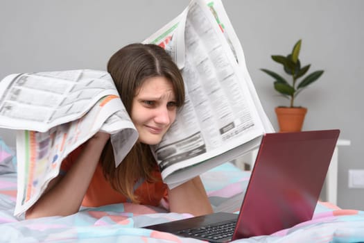 Girl put newspapers to her head and looks at the laptop screen trying to find work