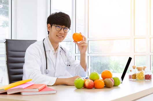 Male nutritionist in white lab coat holding orange fruit in clinic room with fresh variaty fruits and vitamins on his desk. Healthy eating with fruit and vitamin C concept to prevent the flu.