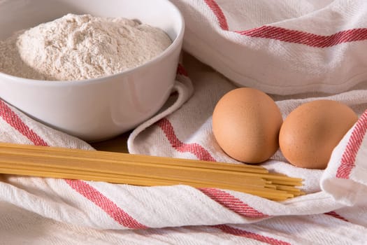 Gourmet close up with a bowl full of flour, spaghetti, and two eggs in white kitchen cloths with red border, on a wooden board-