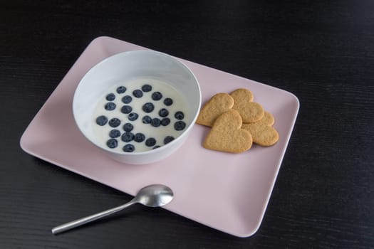 Breakfast. Yogurt with blueberries heart shaped cookies. Ingredients in a pink tray on a black wood table.