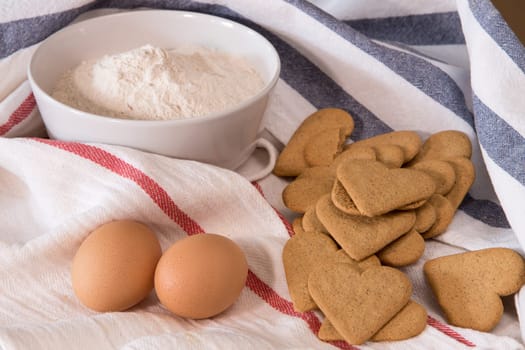 Gourmet close up with a bowl full of flour, heart shape cookies, and two eggs in white kitchen cloths with red and blue border, on a wooden board