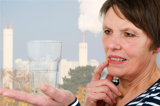 Side head-and-shoulders view of an older woman with a skeptical view of a half-filled glass with water on her right hand standing against an industrial background with a smoking chimney.