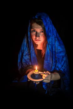 Sad young woman with candle and blue headscarf against black background