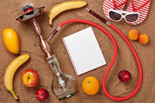 Beach accessories, hookah (shisha), fruit and notebook with blank sheet on sand background. Top view.  Vacation  in warm countries, Beach party and holiday
