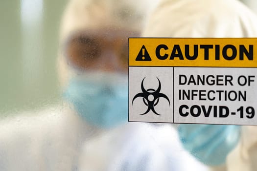 Caution danger Signage of COVID-19 coronavirus in front of Laboratory room with background of Scientists working and researching vaccine for ncov-19 virus for world pandemic situation.