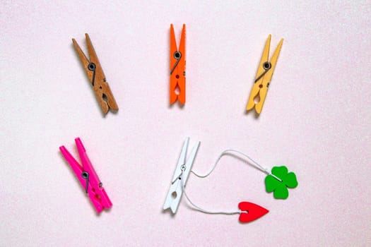 Colorful set of five wooden and plastic clothespins on a light pink glitter sparkle background with space. A red heart and a green lucky clover leaf are attached to the white wooden clip. Topview.