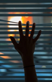 Female hand separating slats of venetian blinds to look through. Silhouette of a hand in the foreground and the blur street in the background
