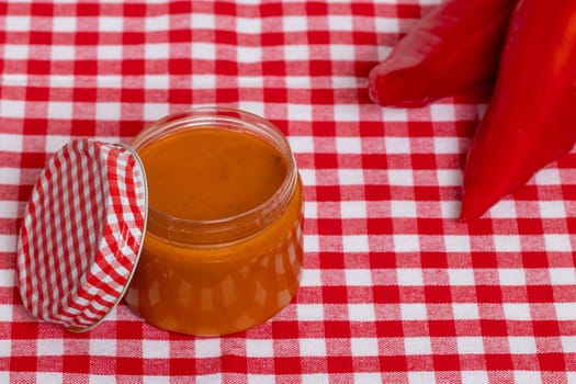 Closeup of a jar with home made classical sauce of fresh paprika with tomatoes, horseradish, garlic, basil and two paprika on a red checkered tablecloth. Template for food product display montage or advertising text. Concept of healthy nutrition. Macro.