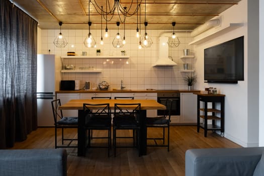 An eat-in kitchen interior design in modern scandinavian style with big wooden table and chairs against light wood floor, bright white walls and furnitures with TV, appliances and hanging light bulbs
