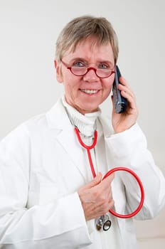 Frontal upper body view of an older woman in a white coat with a stethoscope loosely placed around her neck with a red rubber tube and a wireless phone holding her left ear against a white background, smiling at the camera.