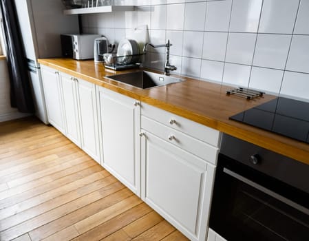 Furniture and appliances: bright white cabinets with wooden countertop, electric cooker, induction hob, faucet, sink and dish rack against hardwood floor and light tile in modern scandinavian kitchen