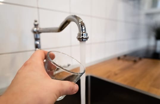 mans hand holding an empty glass nearby a stainless steel kitchen tap. person consider pouring running water from chrome faucet. Potable or Non-potable. Safe to drink or not fit for human consumption
