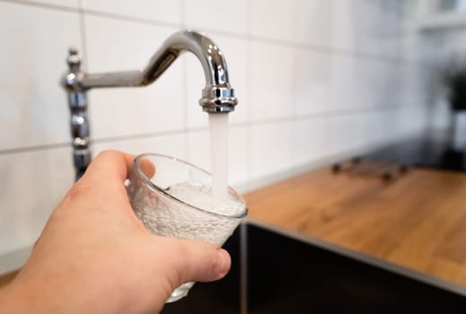 man filling a glass of water from a stainless steel kitchen tap. male's hand pouring water into the glass from chrome faucet to drink running water with air bubbles. potable water and safe to drink