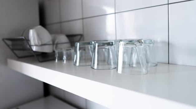 White wooden shelf and budget lightweight dish rack with dishes, cups and glasses against wall tiles at modern scandinavian kitchen. Wide kitchen shelves and wall shelving for storage and display