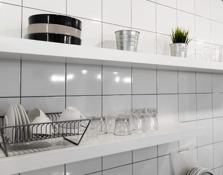 White wooden shelf and budget lightweight dish rack with dishes, cups and glasses against wall tiles at modern scandinavian kitchen. Wide kitchen shelves and wall shelving for storage and display